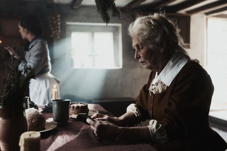 Ann Haltridge sits at a table with a book in her hands. Margaret Spear stands in the background. Dash Productions Services LTD/Antoan Ivanov)