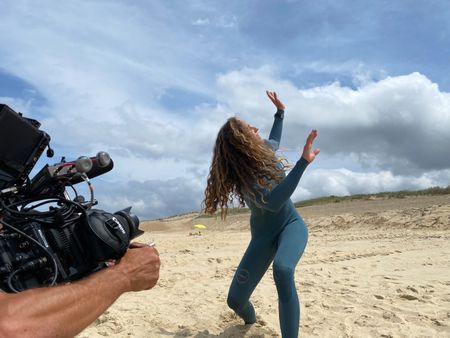 Big wave surfer Justine Dupont, wearing her wetsuit, stretches her body on the beach. DP Alfredo de Juan films her from a distance.   (National Geographic/Gene Gallerano)