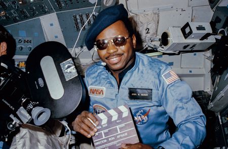 Astronaut Ronald E. McNair, 41-B mission specialist, doubles as "director" for a movie being "produced" aboard the Earth-orbiting Space Shuttle Challenger in February 1984.  McNairís name tag ("Cecil B. McNair"), beret and slate are all humorous props for application of a serious piece of cargo on this eight day flight - the Cinema 360 camera.  Two of the cameras were carried aboard the Challenger to provide a test for motion picture photography in a unique format designed especially for planetarium viewing.  This camera was located in the crew cabin area and a second was stowed in a getaway special (GAS) canister in the payload bay.  (credit: NASA)