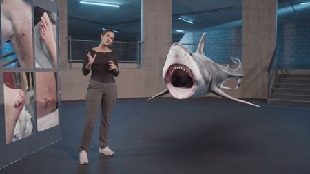 Dr. Diva Amon speaking to camera next to GFX 'Coral' information board that is displaying the real injuries that Peter Banculli, contributor, sustained to his foot from a shark bite and whilst a GFX Great White shark shows off its impressive set of teeth in the background. (National Geographic)