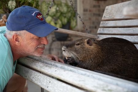 Denny smiling at Nutria the beaver on a porch swing. (Big Wave Productions)