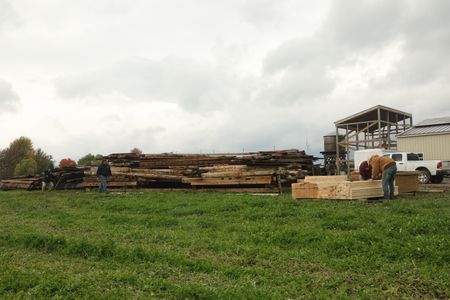 Charles Pol walks towards Andrew Hutton and Seth Doble, who are measuring pieces of lumber they will be using to build a box around the old barn they transported, as a crew member films. (National Geographic)