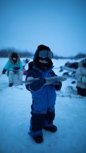 Sabastian Hailstone holds up a fish while his mother, grandmother and aunt ice fish next to him. (BBC Studios Reality Productions, LLC/Pedro Delbrey)