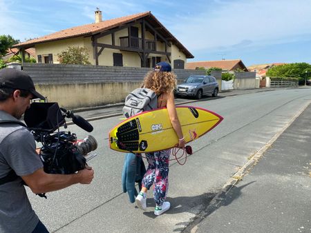 Big wave surfer Justine Dupont walks on the streets with her board under an arm. DP Alfredo de Juan follows her filming with a camera.   (National Geographic/Gene Gallerano)