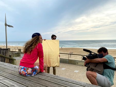 Big wave surfer Justine Dupont sits on steps on the boardwalk next to the beach. DP Alfredo de Juan films from below and AC Florian Morel holds the light reflector.  (National Geographic/Gene Gallerano)