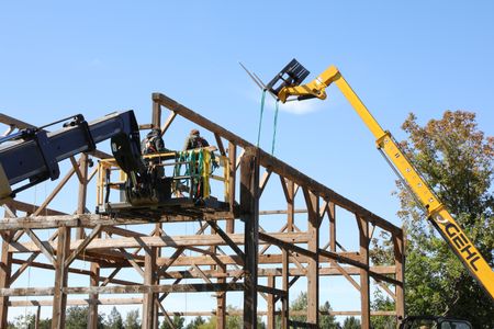 Charles Pol and Ben Reinhold stand on a boom lift above the old barn as a telehandler lifts a piece of the barn's frame. (National Geographic)