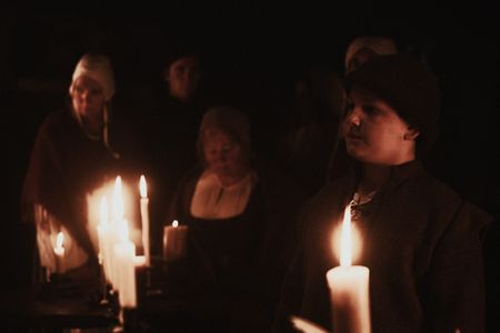 Mats stands in a candle-lit room and villagers observe in the background.(Dash Productions Services LTD/Antoan Ivanov)