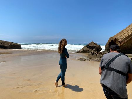 Big wave surfer Justine Dupont, wearing her wetsuit, walks on the beach with her feet in the water. DP Alfredo de Juan follows her, filming.   (National Geographic/Gene Gallerano)