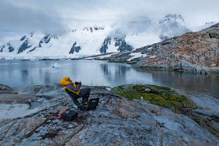 Bertie Gregory flies a drone sitting on a rock against the backdrop of a steep snowy mountain range.  (credit: National Geographic/Bertie Gregory)