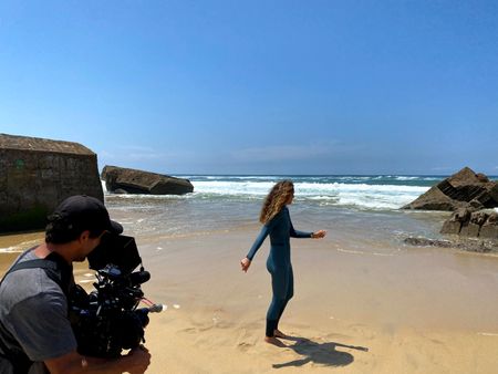 Big wave surfer Justine Dupont, wearing her wetsuit, walks on the beach with her feet in the water. DP Alfredo de Juan follows her, filming.   (National Geographic/Gene Gallerano)