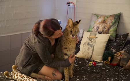 Clare sitting on the bed while hugging a serval. (Big Wave Productions)