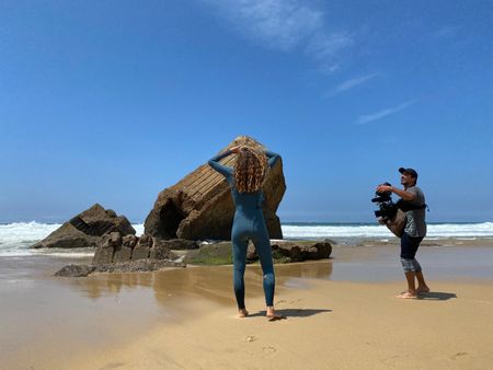Big wave surfer Justine Dupont, wearing her wetsuit, stretches on the beach, with her feet in the water and hands grabing her hair. DP Alfredo de Juan follows her, filming.  (National Geographic/Gene Gallerano)