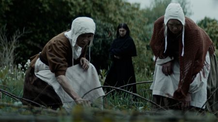 Two village women are kneeling in a field foraging. A villager in dark clothing stands in the background. (Dash Pictures/Samuel Purcell, Oliver Watts)