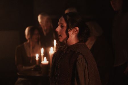 Gertrude stands in a candle-lit room with villagers in the background. (Dash Productions Services LTD/Antoan Ivanov)