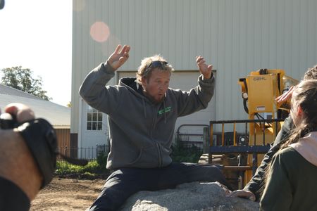 Ben Reinhold sits on the large centerpiece rock and makes hand gestures, while Charles Pol and Beth Pol watch and a crew member films. (National Geographic)