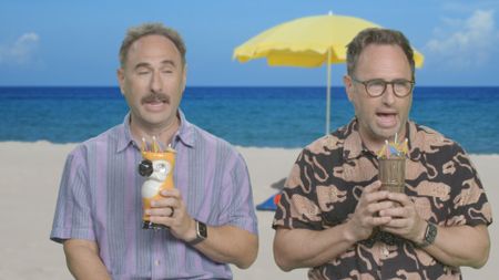 Comedians Randy and Jason Sklar drinking tropical drinks with a yellow beach umbrella on a sandy tropical paradise in Thailand in the background. (National Geographic)