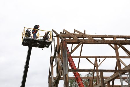 Charles Pol and Ben Reinhold on a lift above the leftover framework from the old barn they are taking down, while a crew member films them. (National Geographic)