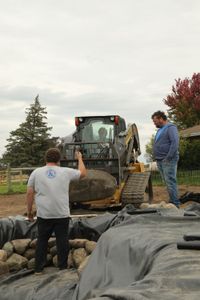 Andrew Hutton guides Ben Reinhold, in a track loader, as he transports large rocks for the farm's new pond, while Charles Pol watches. (National Geographic)