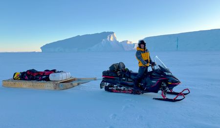 Bertie Gregory on a snowmobile with a sledge loaded with camera equipment on the sea ice, with icebergs in the background. (credit: National Geographic/Heather Cruickshank)