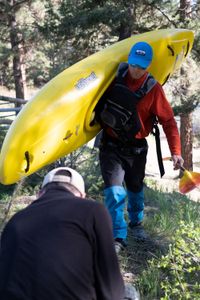 Ben Stookesberry, wearing kayaking outfit, hat and sunglasses, walks in the woods carrying his kayak and his paddle.  DP Ross McDonnell is crouching in front of him getting the shot.  (National Geographic/Harrison Gayton)