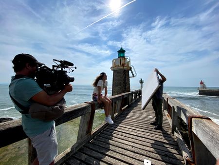 Big wave surfer Justine Dupont sits on the pier's handrail admiring the sea, as DP Alfonso de Juan films her and Fixer Mathias Bertoux holds a light reflector.  (National Geographic/Gene Gallerano)