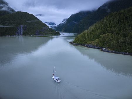 Aerial view of the sail boat passing through the Inside Passage, surrounded by mountains and trees. Alex Honnold and Tommy Caldwell are standing on the prow of the ship.  (National Geographic/Renan Ozturk )