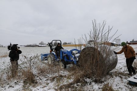A crew member films Charles Pol driving a tractor while Beth Pol pushes a bale of hay into the bucket to transport it to the bee hives so it can block strong winter winds from harming the hives. (National Geographic)