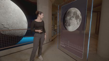 Dr. Diva Amon in the shark lab studio while standing next to GFX 'Coral' information board which is displaying an image of the moon. (National Geographic)