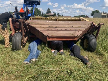 A crew member films Dr. Pol, Charles Pol, and Ben Reinhold crawling under the mower conditioner to pull the hay plugging up the blades. (National Geographic)